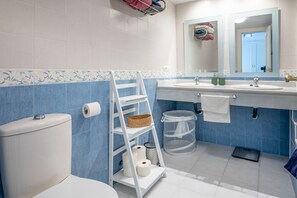 Een badkamer met toilet, wastafel en spiegel. Er is een witte plank met handdoeken en een mand.
