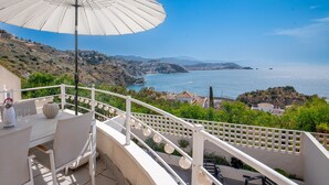 Une terrasse avec une table, des chaises et un parasol, d’où l’on peut admirer un paysage côtier avec des maisons et la mer.