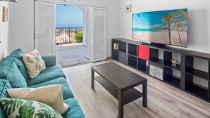 A living room with a teal sofa, a coffee table, a TV on a black stand, and a balcony with a view of the sea.