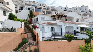 A white building with a staircase leading to a balcony, surrounded by greenery and other white buildings.