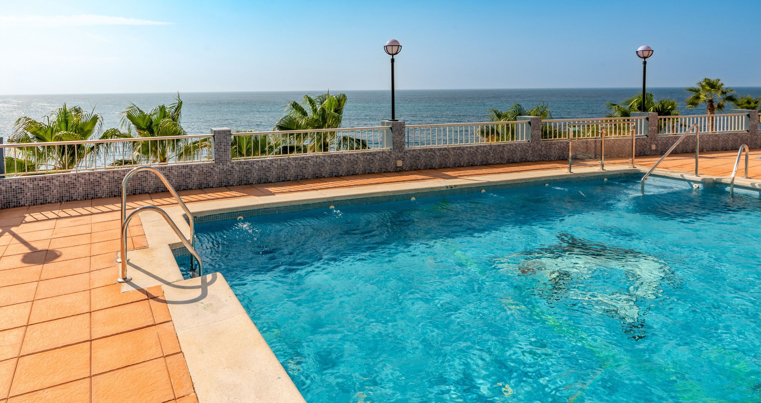Piscina con vistas al océano, rodeada de palmeras y un solárium con baldosas.