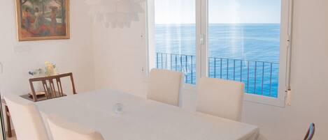 A dining area with a table and chairs, a painting on the wall, and a view of the sea through large windows.
