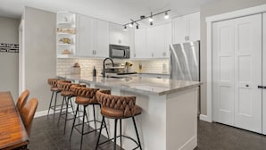 A modern kitchen with white cabinets, a central island with stools, a microwave, and a stainless steel refrigerator. - Water`s Edge | Pool | Hot Tub | Pets | Ground LVL (Invermere)