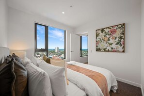 A modern hotel room with a large bed, a sofa, a desk, and a view of the cityscape through large windows.