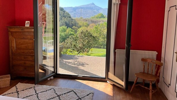 Ein Zimmer mit roter Wand, einem hölzernen Schminktisch, einem Stuhl und einer Glastür, die auf einen Balkon mit Bergblick führt.
