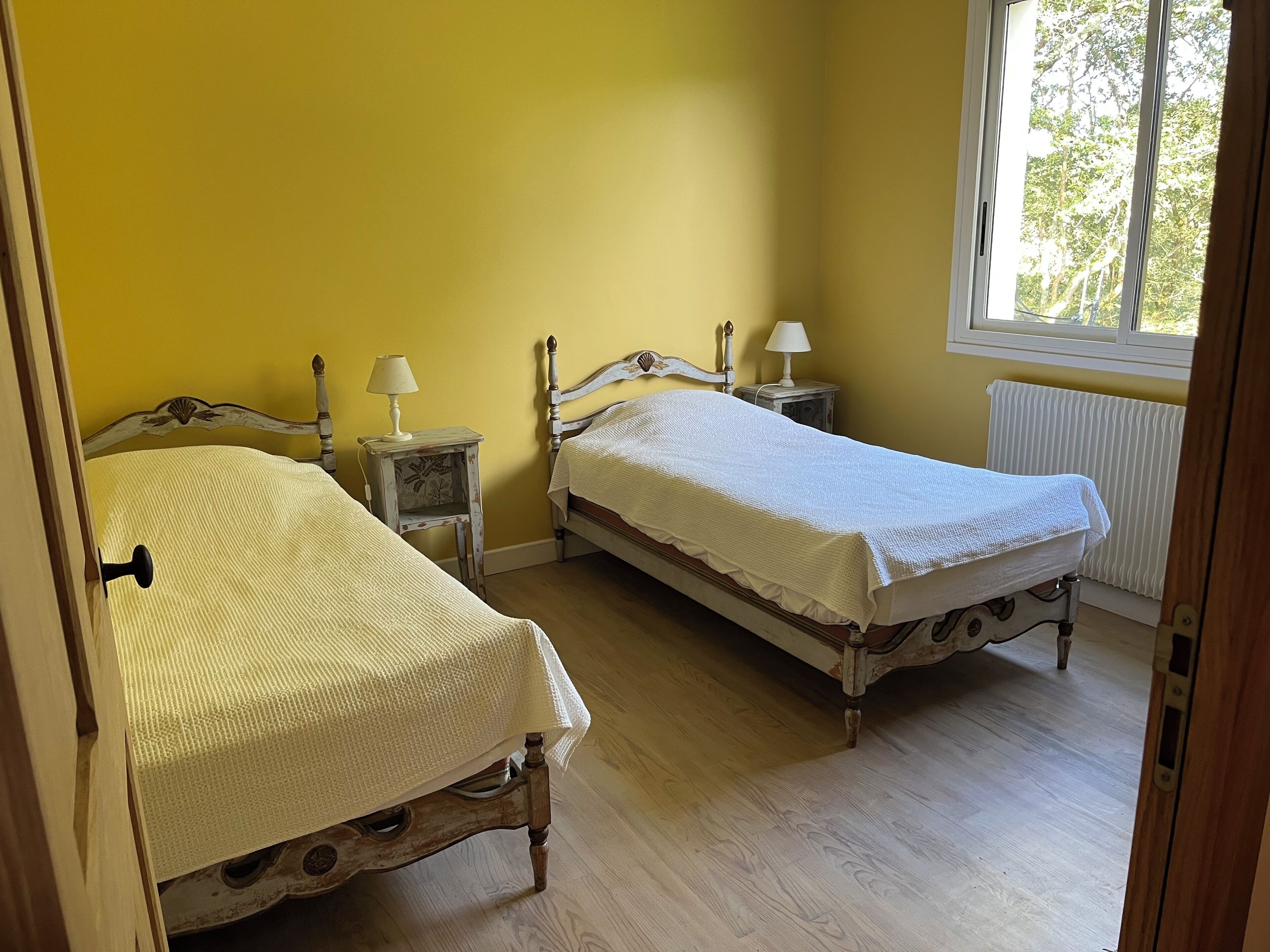 Une chambre à coucher avec deux lits en bois, chacun recouvert d’une courtepointe blanche et agrémenté d’une petite table de chevet avec une lampe. Les murs de la pièce sont peints en jaune, il y a une fenêtre donnant sur des arbres et un radiateur.