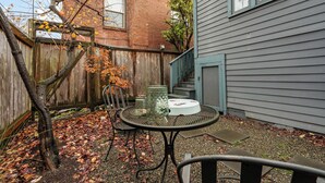 Un patio con una mesa y sillas de metal redondas, una valla de madera y un edificio de ladrillo al fondo.