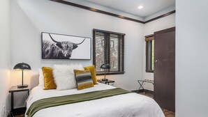 Un dormitorio con cama, mesita de noche, lámpara, ventana con persianas, puerta y un cuadro colgado en la pared.