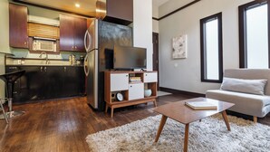 Una cocina moderna con armarios de madera oscura, un refrigerador de acero inoxidable y un horno empotrado. Hay una mesa de centro de madera con un libro y una alfombra mullida. Un sofá gris se encuentra en la sala de estar, junto a un ventanal grande.
