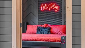 A cozy room with a bed, a neon sign reading "Let's Play," and a black textured wall.