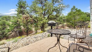 Eine Terrasse im Freien mit einem runden Metalltisch und Stühlen, einem Grill und Blick auf eine gebirgige Landschaft.