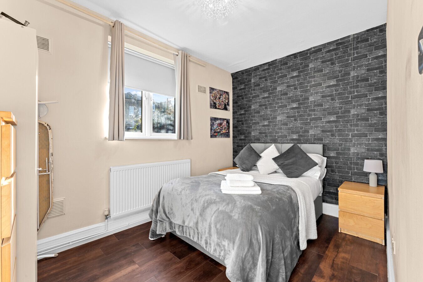 A bedroom with a bed, a radiator, a window with curtains, a brick wall, and a nightstand with a lamp.