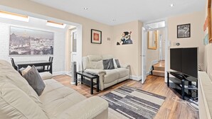 Una sala de estar moderna con un sofá, una mesa de centro y un televisor. Hay un ventanal con vista a un paisaje urbano, una puerta que conduce a otra habitación y un cuadro en la pared.