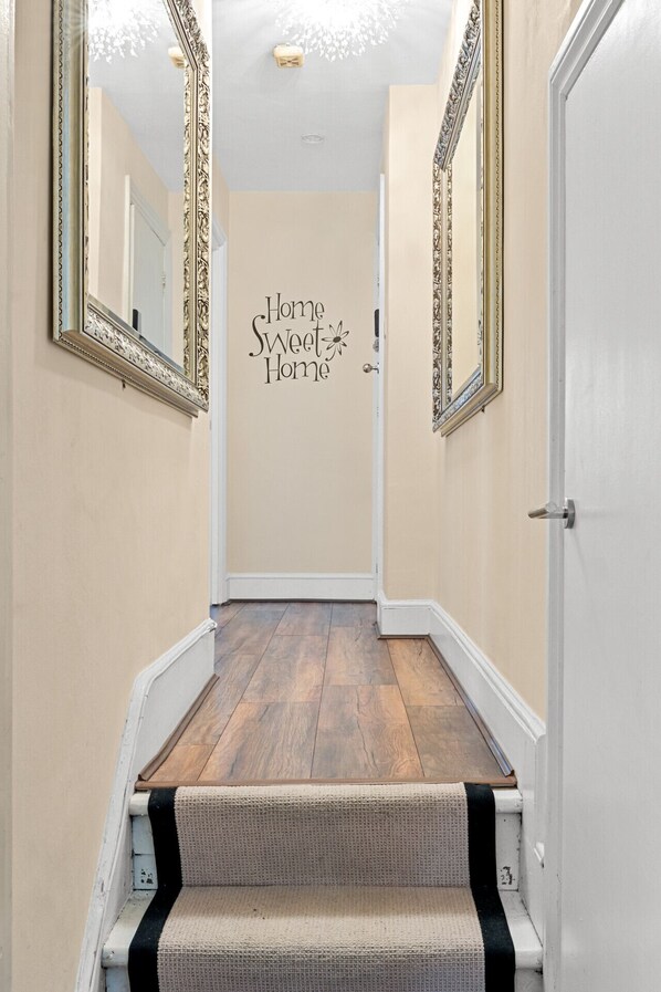 A hallway with a wooden floor, a carpeted staircase, and a wall with the text "Home Sweet Home".