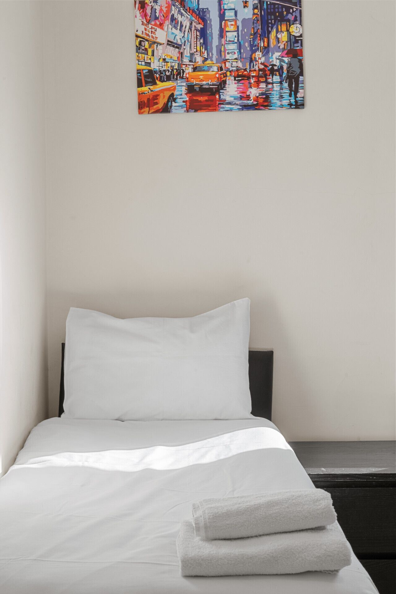 Een net opgemaakt bed met witte lakens en een opgevouwen handdoek, een effen muur en een kleurrijk schilderij van een levendige straat boven het bed.