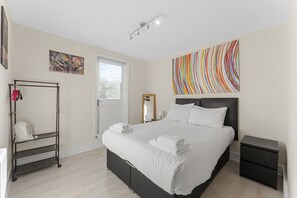 A modern bedroom with a large bed, a standing fan, a window, and a colorful abstract painting.