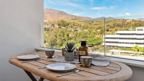 Ein Holztisch mit Tellern, Besteck, einer Tasse, einem kleinen Topfpflanzen und einer Flasche auf einem Balkon mit Blick auf eine Berglandschaft und Gebäude.