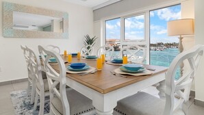 Un coin repas avec une table en bois dressée pour quatre personnes, des chaises blanches, une grande fenêtre donnant sur la mer et un miroir accroché au mur.