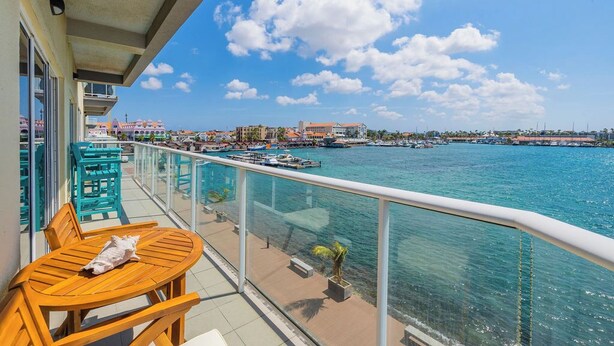 Un balcon doté de mobilier en bois, donnant sur une marina avec des bateaux et des bâtiments en arrière-plan.