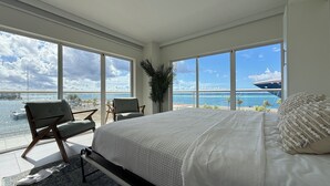 Une chambre spacieuse avec un grand lit, un coin salon avec des fauteuils et une vue sur l’océan et un navire.