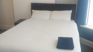Una cama bien hecha con sábanas blancas y dos almohadas, una toalla azul oscuro doblada al pie de la cama, y una mesita de noche con una lámpara a la izquierda.