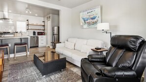 Una sala de estar moderna con un sofá blanco, un sillón de cuero negro y una mesa de centro de vidrio. Incluye una zona de cocina con microondas, cafetera y campana extractora de acero inoxidable. Un cuadro con barcos adorna la pared, y una lámpara está sobre una mesita auxiliar.