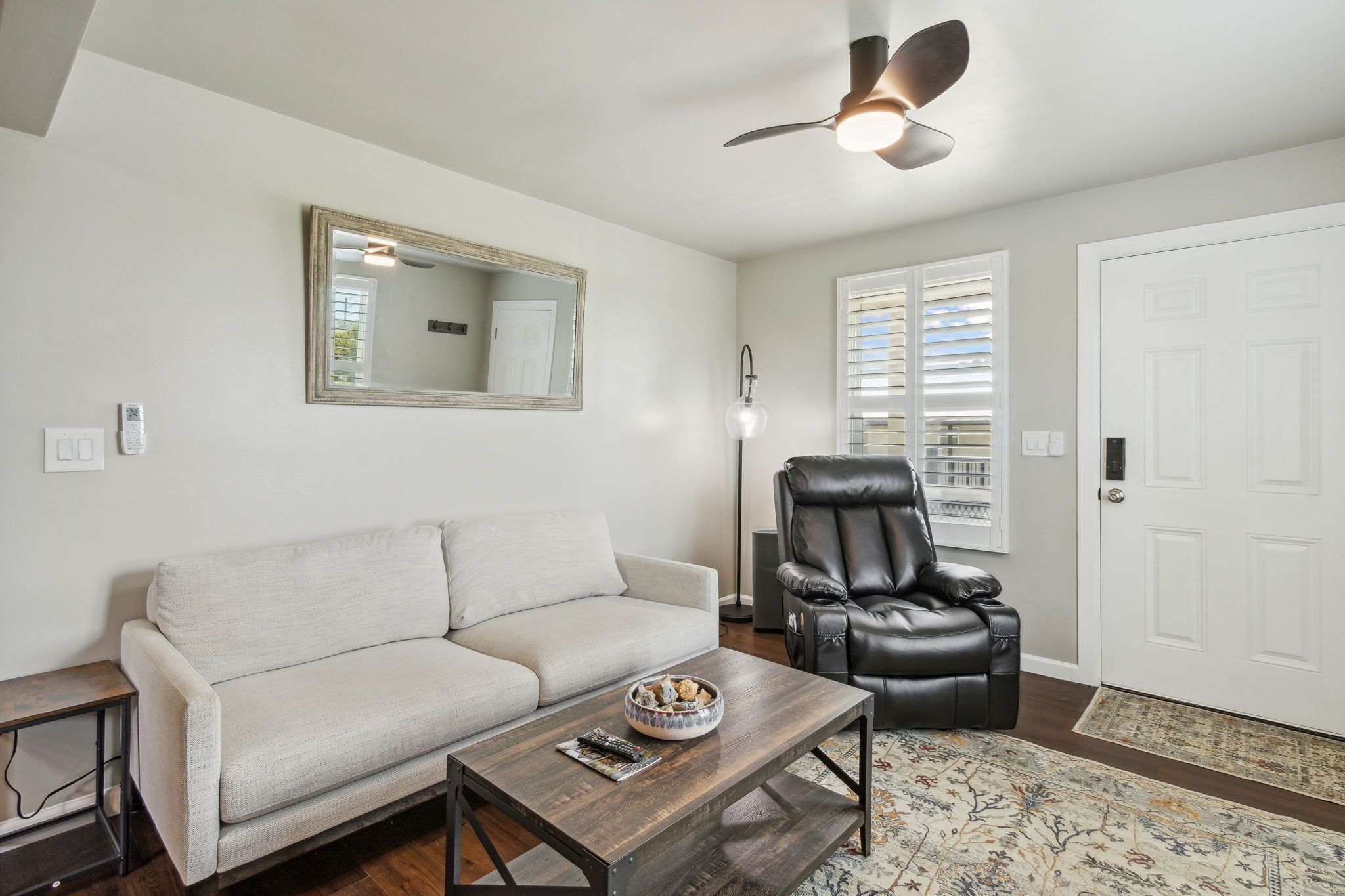 A living room with a sofa, a coffee table, a black leather chair, a ceiling fan, a mirror, and a door with blinds.