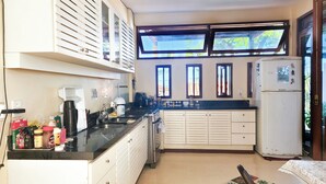 A compact kitchen with a gas stove, sink, and a refrigerator. There are multiple windows providing natural light, and the kitchen has a simple, functional design. - Casa Aberta (Itacaré)