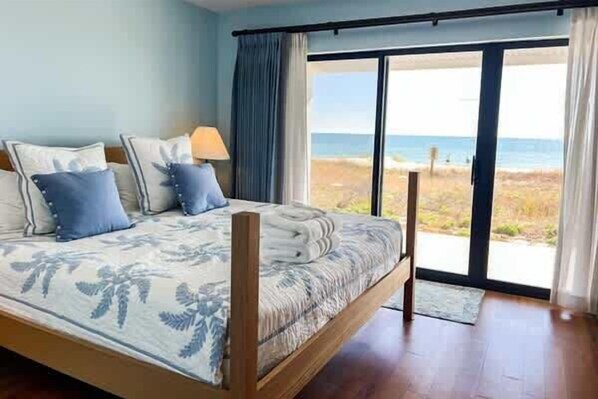 Un dormitorio con cama, una mesita de noche, una lámpara y vista a la playa a través de un ventanal corredero.