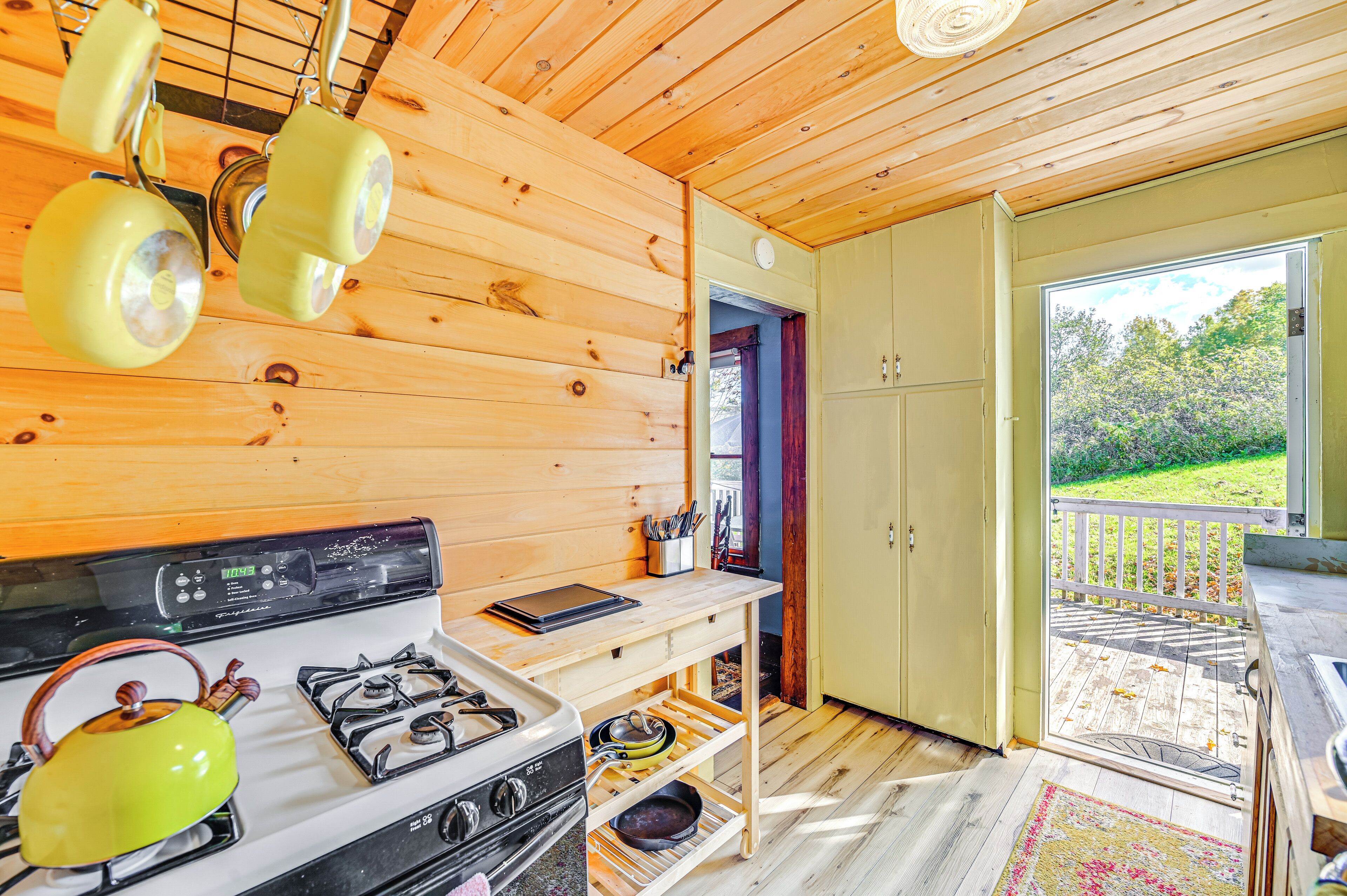 L’intérieur d’une cabane en bois comprend un coin cuisine équipé d’un poêle, d’un évier et de quelques ustensiles de cuisine suspendus. On y trouve également une étagère en bois où sont rangés des ustensiles de cuisine, ainsi qu’une petite table. Une porte donne accès à une terrasse extérieure dotée d’une rambarde.