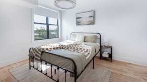 Een moderne slaapkamer met een groot raam, een bed met metalen frame, een nachtkastje met lamp en een aan de muur hangend kunstwerk.