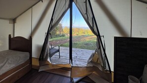 A tented room with a bed, a chair, and a view of a grassy field with trees.