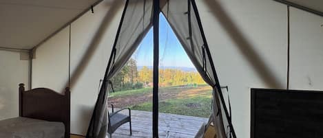 A tented room with a bed, a chair, and a view of a grassy field with trees.
