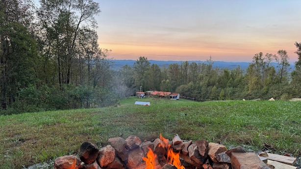 A campfire surrounded by rocks and logs, with a view of a house and trees in the background.