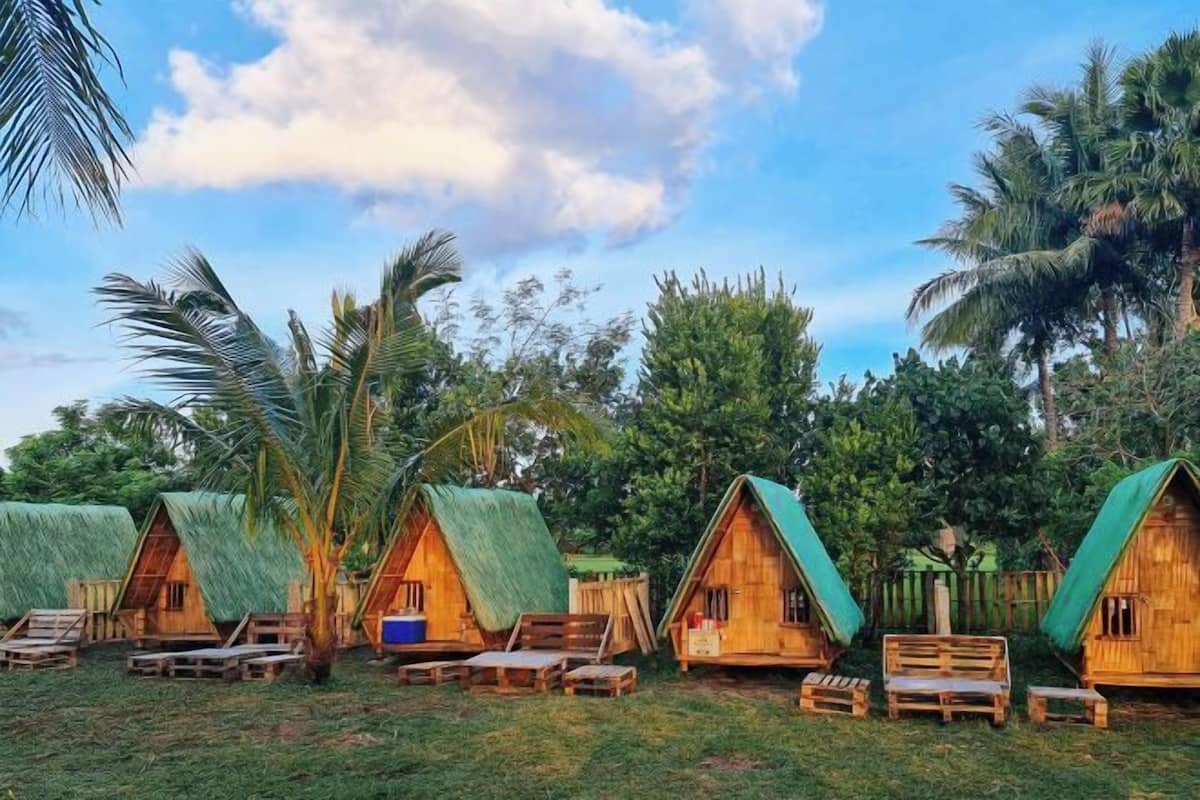 A row of wooden huts with thatched roofs, each with a bench and a small table.