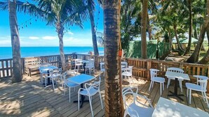 Une terrasse extérieure avec des tables et des chaises blanches, des palmiers et une vue sur l’océan.