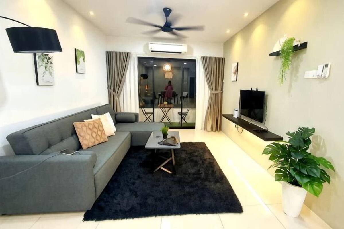 A modern living room with a grey sofa, a black rug, a ceiling fan, a flat-screen TV, and a potted plant.