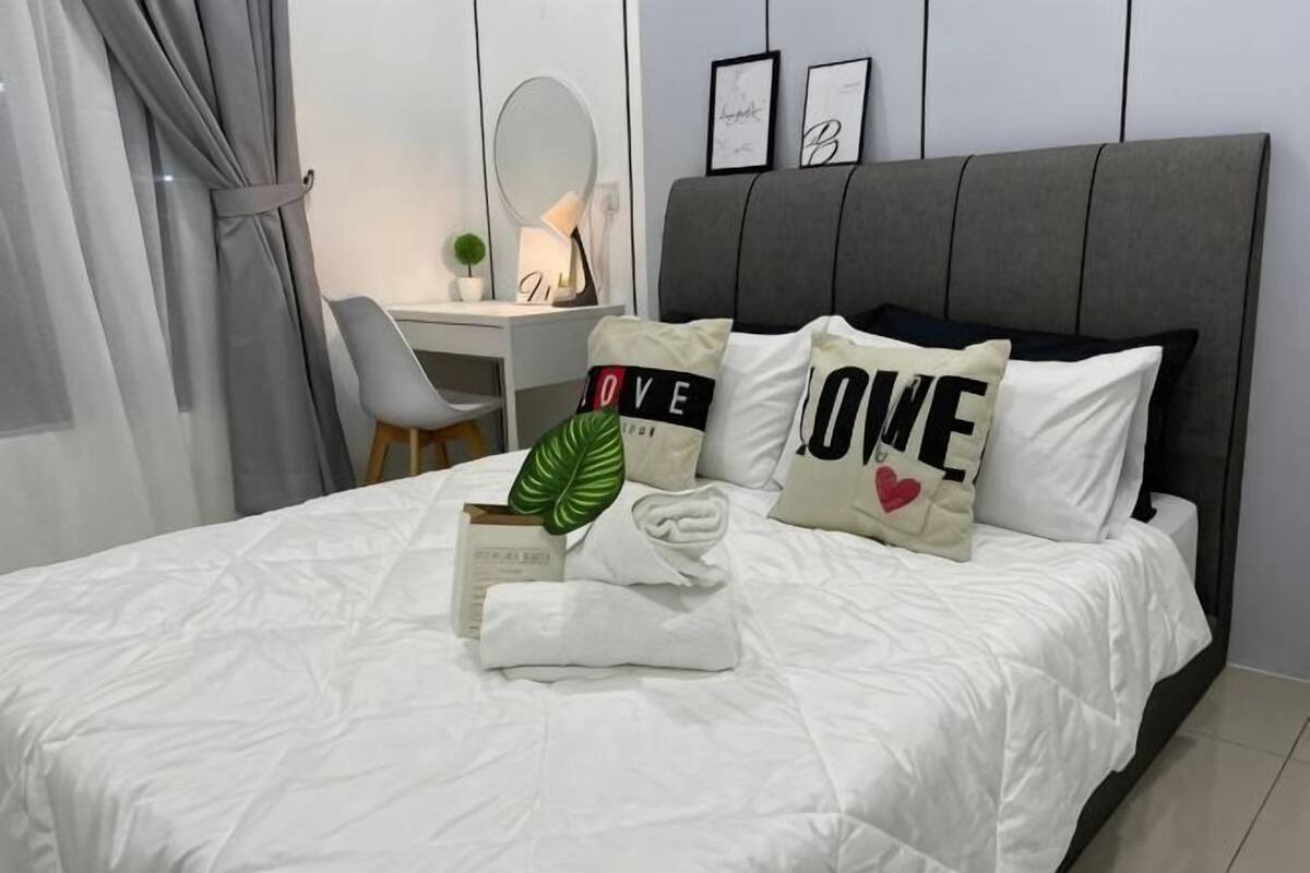 A neatly made bed with pillows, a bedside table with a lamp, a chair, and a desk with a computer.