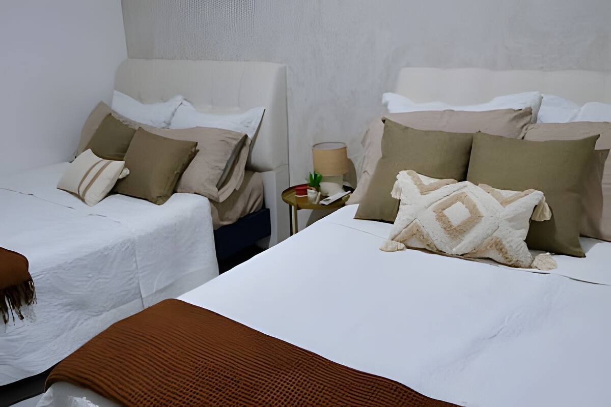 Quarto de hotel com duas camas de solteiro, cada uma com um edredom branco e várias almofadas. Há uma pequena mesa redonda com um abajur e um objeto vermelho em cima.