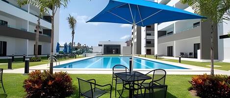 A modern pool area with a blue umbrella, surrounded by well-maintained grass and plants, and adjacent to a multi-story building.