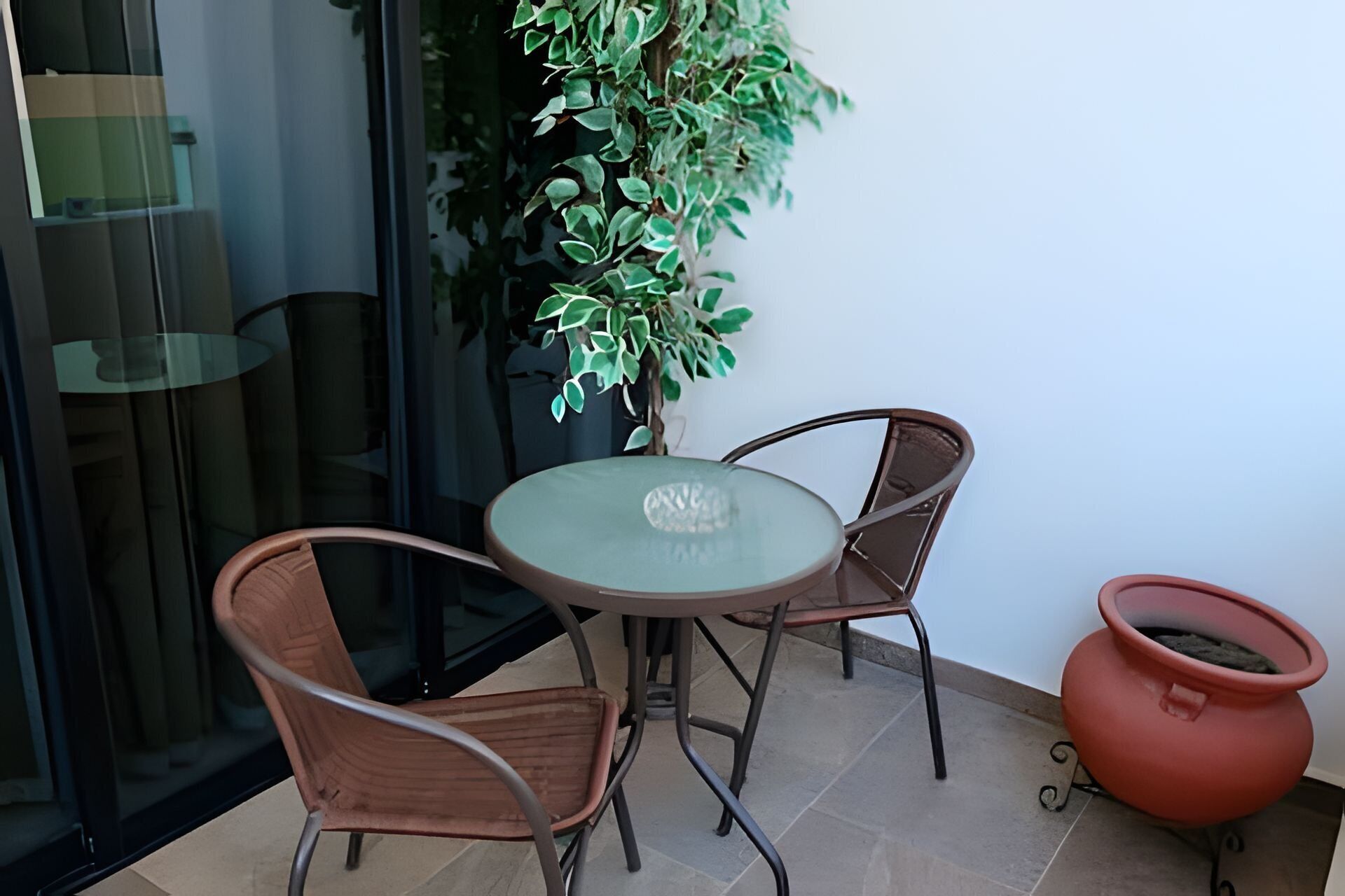Un pequeño espacio exterior con una mesa de vidrio y dos sillas, una planta en maceta y una maceta roja.