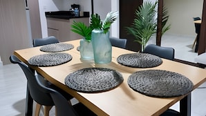 A dining table with six woven placemats, a glass vase with green plants, and a modern kitchen in the background. - Marina cerritos Torre 1 (Mazatlán)