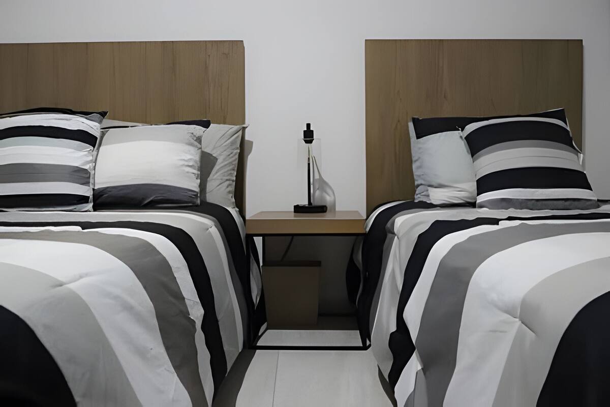 Quarto de hotel com duas camas, cada uma com roupa de cama listrada em preto e branco. Há uma mesa de cabeceira com abajur entre as camas.