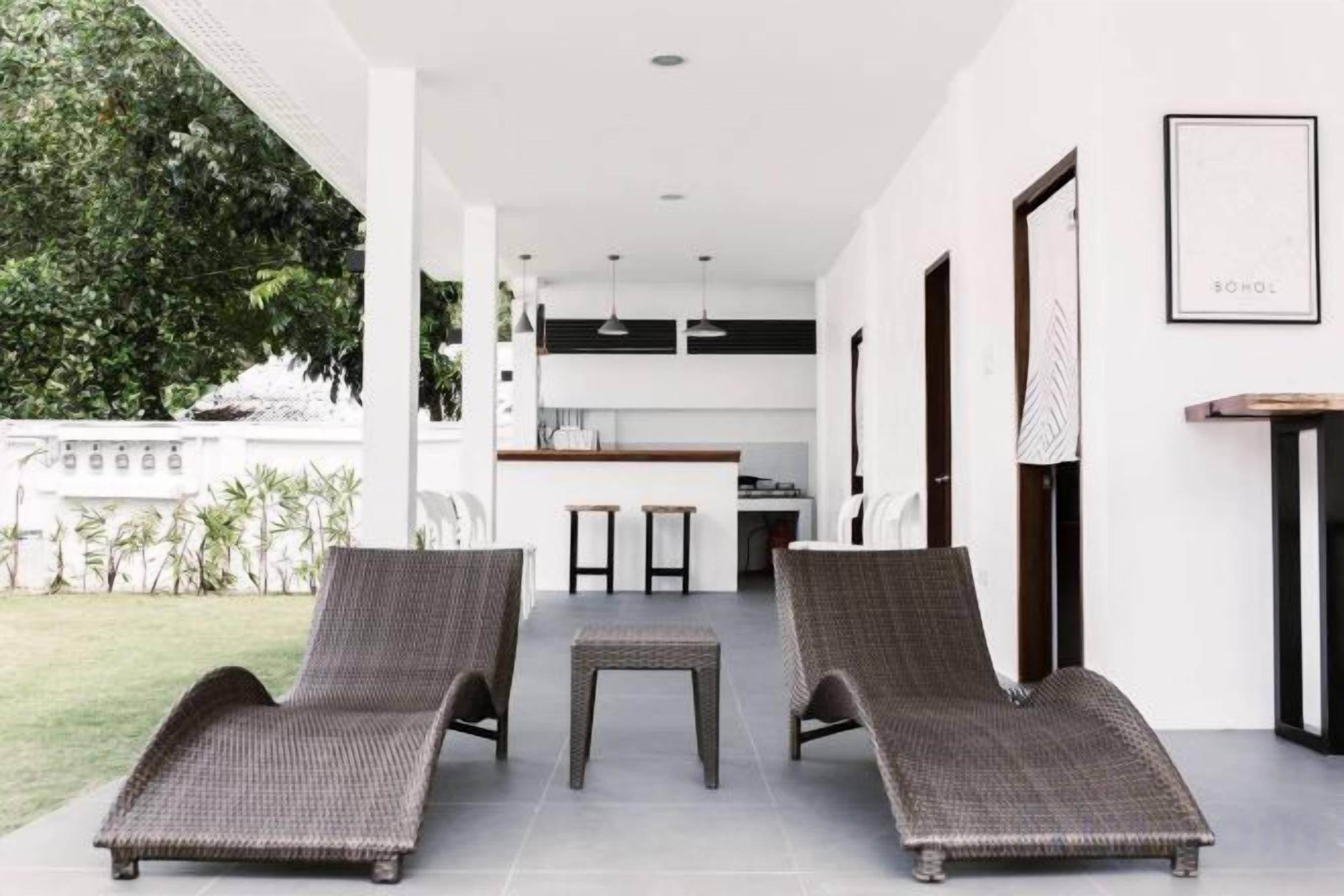 Un espace de détente extérieur moderne, avec deux fauteuils en osier et une petite table, une cuisine en arrière-plan et une œuvre d’art encadrée accrochée au mur.