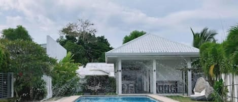 A swimming pool with a covered pavilion, surrounded by greenery and a clear sky.
