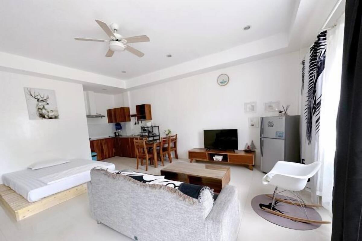 A modern living room with a sofa, a TV, a dining area, and a kitchen in the background.