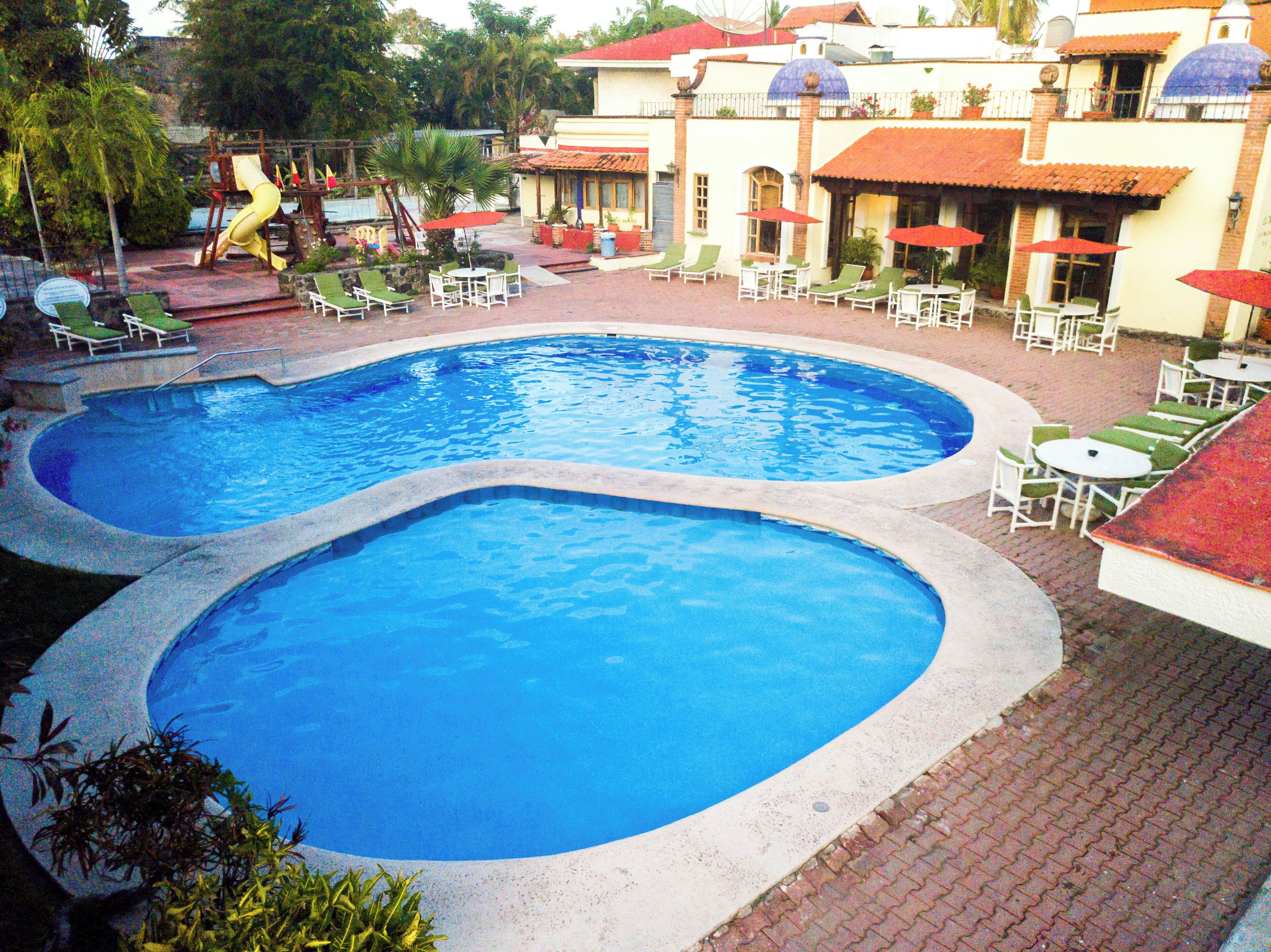 A swimming pool with two distinct sections, surrounded by lounge chairs and umbrellas, and a building with a red-tiled roof in the background.