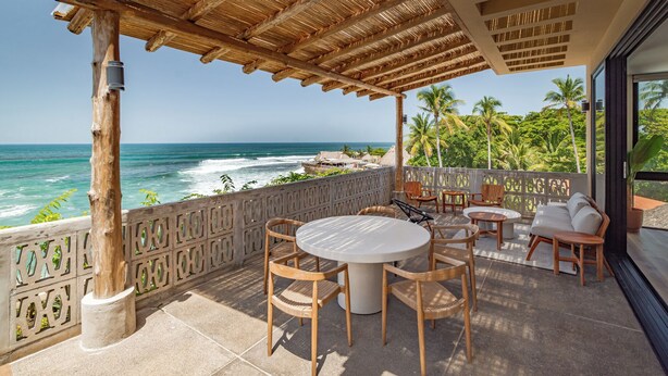 A covered outdoor seating area with a view of the ocean, featuring a round table and chairs, a wooden pergola, and a decorative fence. - Wave House (Chiltiupan)