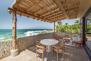 Un espace extérieur aménagé, abrité et doté de sièges, offrant une vue sur l’océan, avec une table ronde et des chaises, une pergola en bois et une clôture décorative.