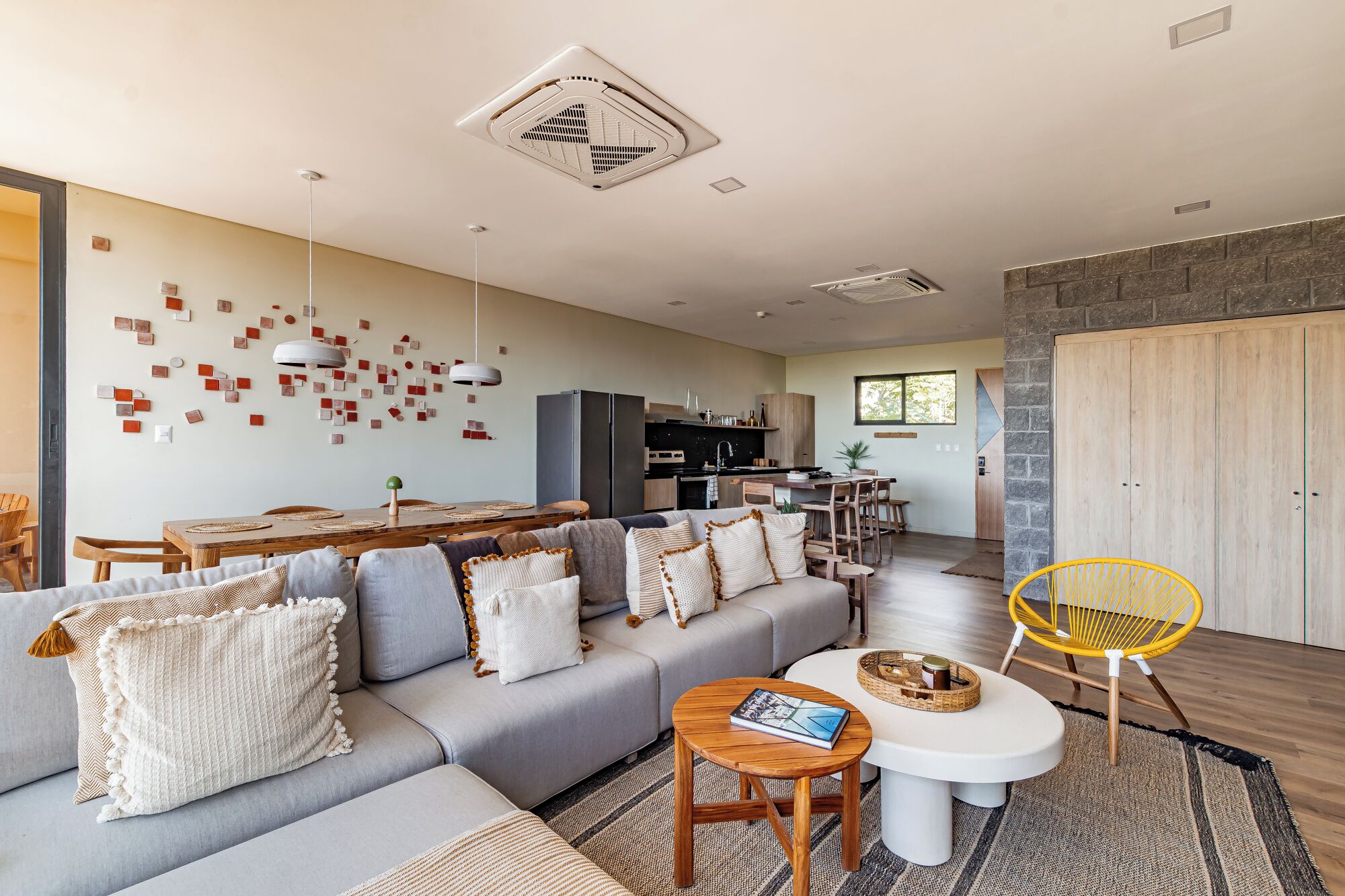 A modern living room with a grey sofa, wooden coffee table, and yellow chair. There is a kitchen area with a fridge and cabinets.